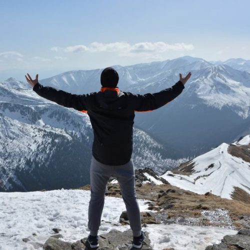 A man stands on a snowy mountain peak with arms raised, embracing the breathtaking view.