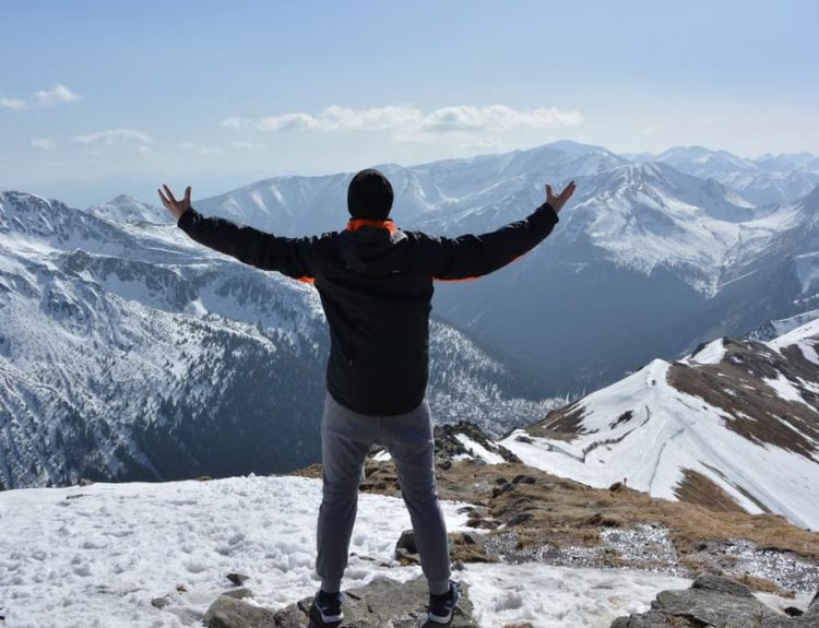 A man stands on a snowy mountain peak with arms raised, embracing the breathtaking view.